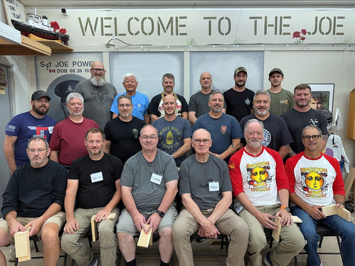 A group of 18 men posing together in an indoor workshop or shop environment, with a large sign reading 'WELCOME TO THE JOE' on the wall behind them. Several men are wearing t-shirts with 'Sgt.